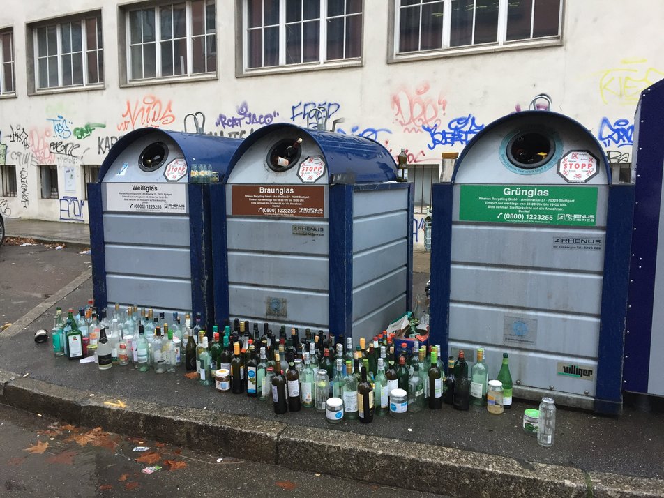 Drei Glascontainer, vor denen Leergut steht